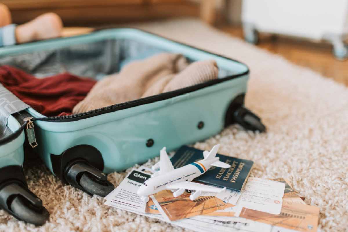 Sopra ad un tappeto bianco c'è una valigia celeste aperta con davanti dei documenti di viaggio e un aeroplanino giocattolo